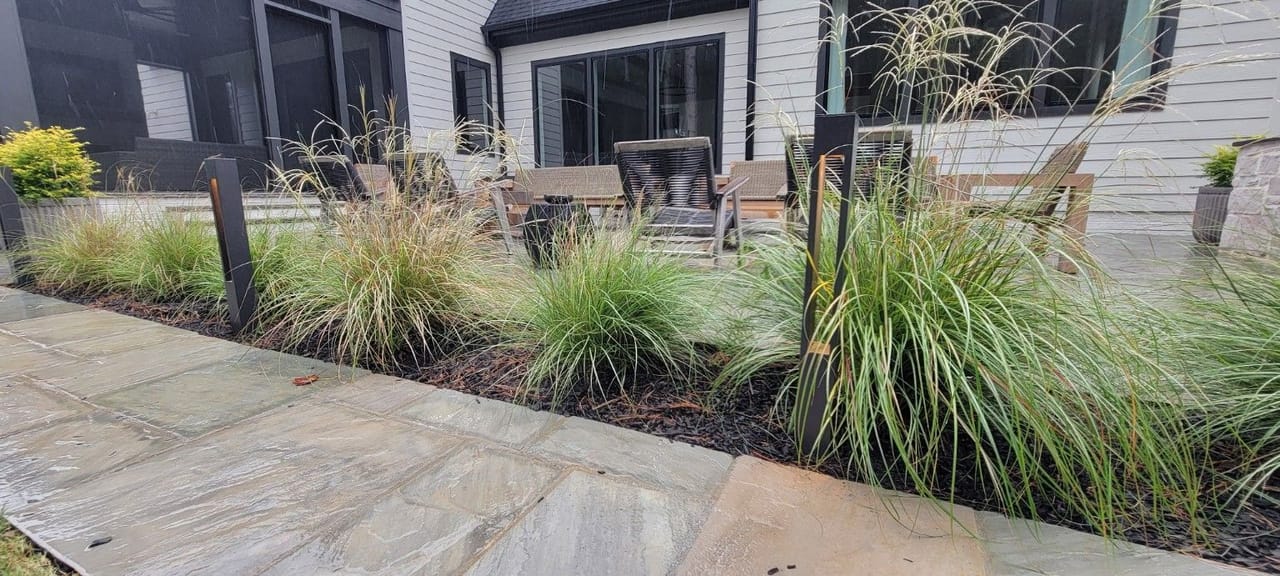 Black bollard path lights in an ornamental grass bed along a flagstone patio with seating, rainy daytime in Wake County