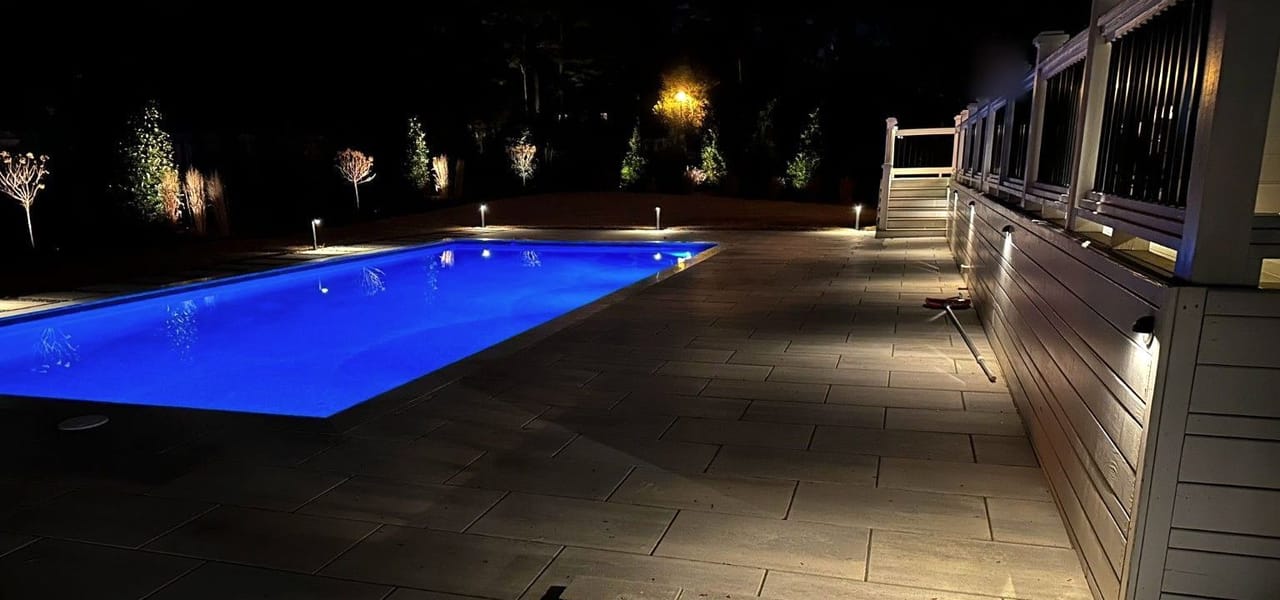 Blue-lit pool with path lights, up-lit trees and deck wall lights on a paver patio at night in Wake County