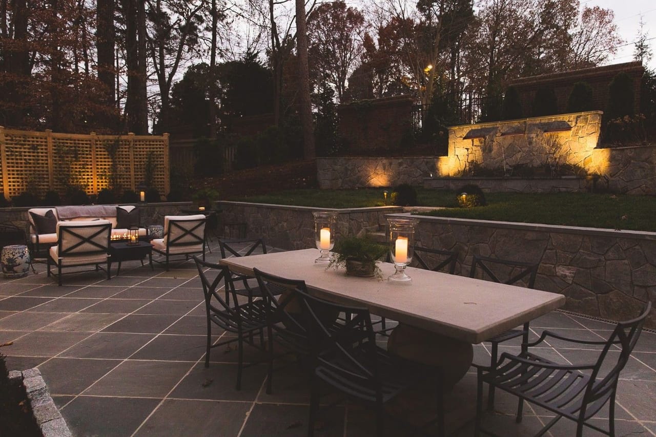 Bluestone patio with dining table and fire table, up-lit stone water wall and lattice fence at dusk in Wake County