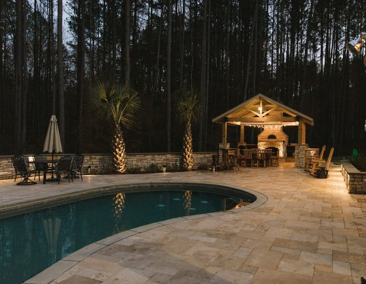 Travertine pool deck with up-lit palms, seat-wall lights and covered pavilion with fireplace at dusk in Wake County