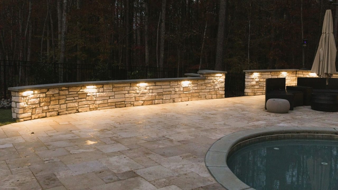 Stacked-stone seat wall with under-cap lights glowing over a travertine pool deck at dusk in Wake County