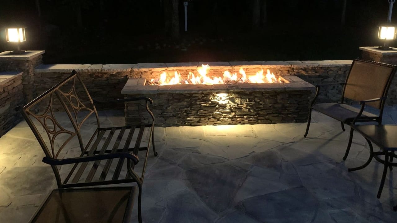 Linear gas fire pit in a stacked-stone wall with an under-cap light on a flagstone patio at dusk in Wake County