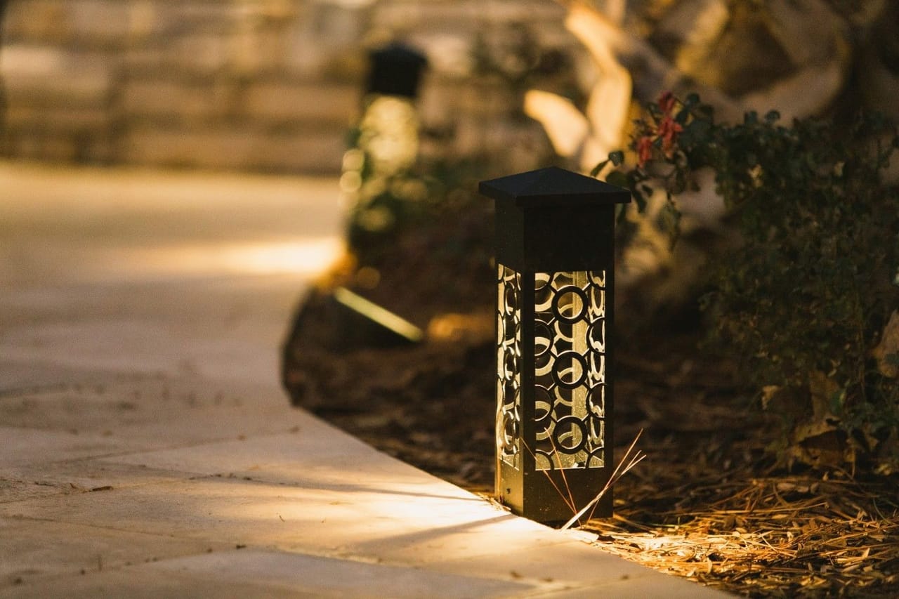 Close-up of a black pierced-pattern path light glowing beside a pale stone walkway and pine-straw bed at night in Wake County