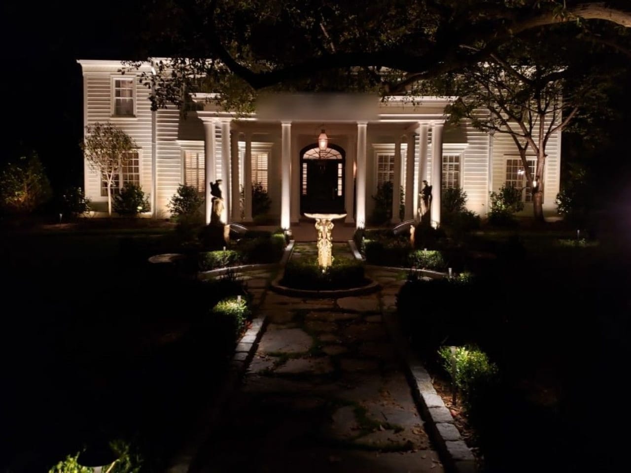 White colonial home with uplit columns and a stone walkway lined with path lights leading to a lit fountain at night in Wake County