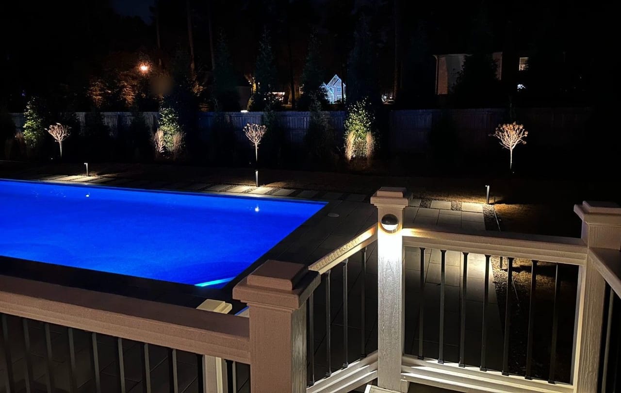 Blue-lit pool with path lights along the paver deck and uplit ornamental trees by the fence, seen from a lit deck rail at night in Wake County
